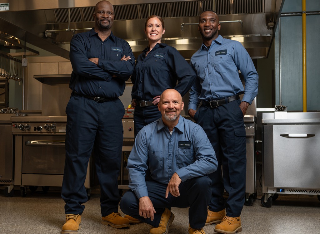 Team of four commercial kitchen service technicians in uniform posing together in a restaurant kitchen