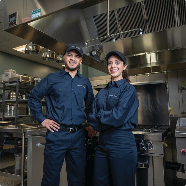 Two commercial kitchen service technicians in uniform standing among cooking equipment