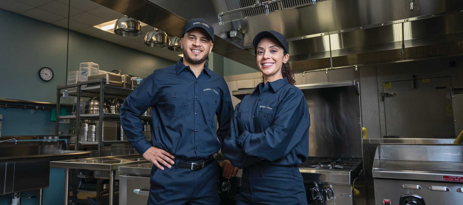 Two commercial kitchen service technicians in uniform standing among cooking equipment