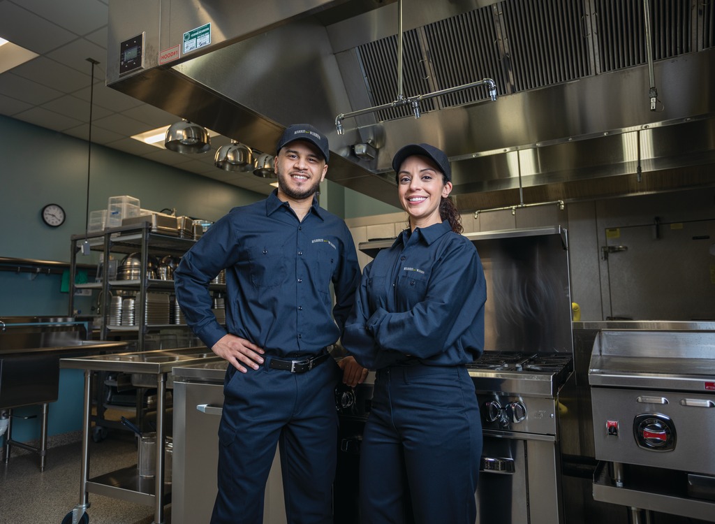 Two commercial kitchen service technicians in uniform standing among cooking equipment