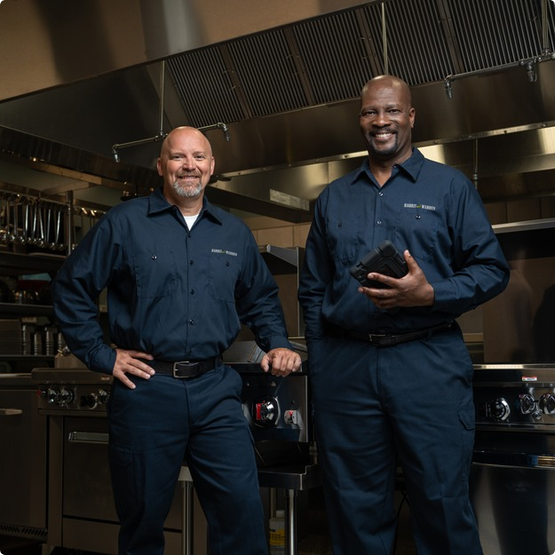 Two commercial kitchen service technicians in uniform standing in a restaurant kitchen