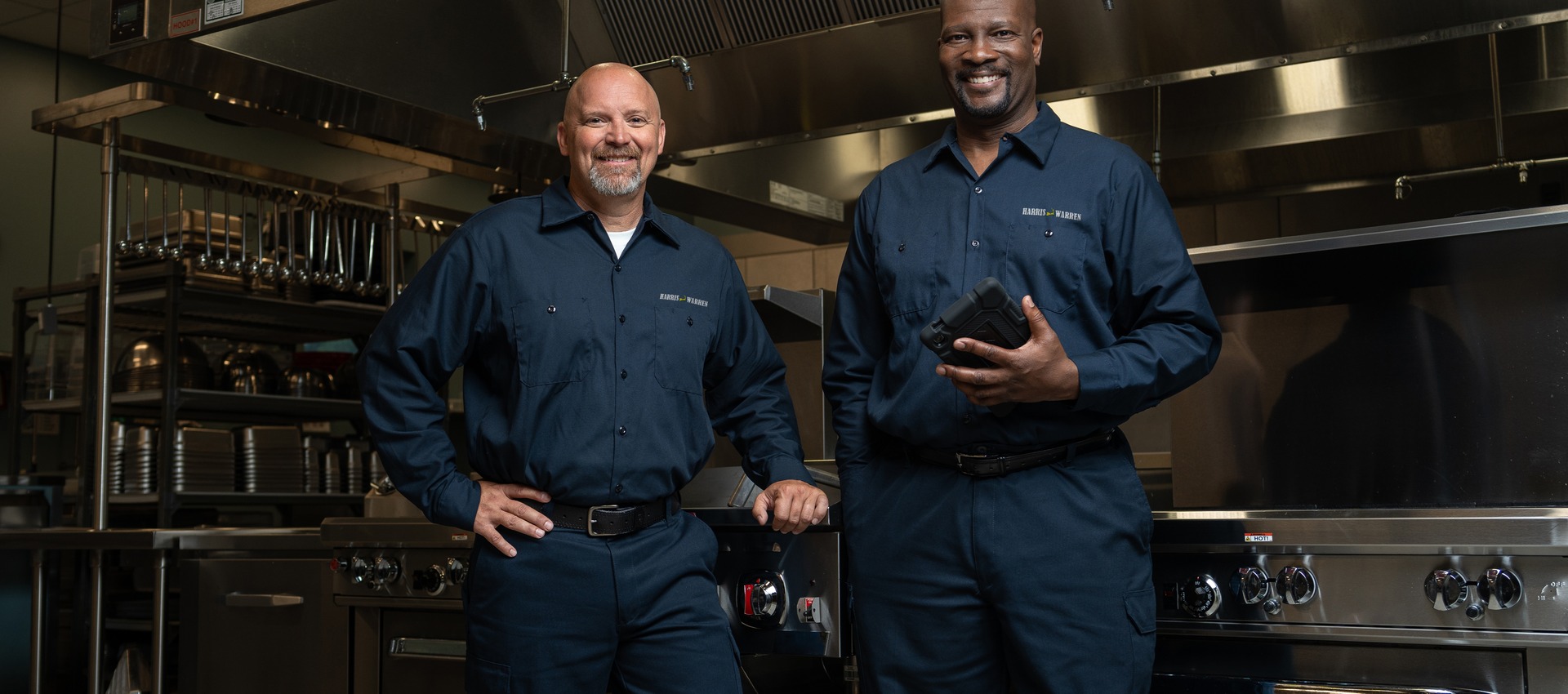 Two commercial kitchen service technicians in uniform standing in a restaurant kitchen