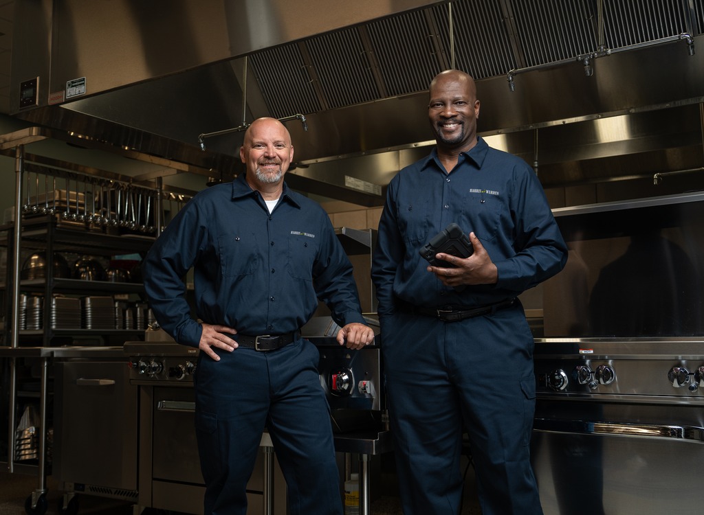 Two commercial kitchen service technicians in uniform standing in a restaurant kitchen